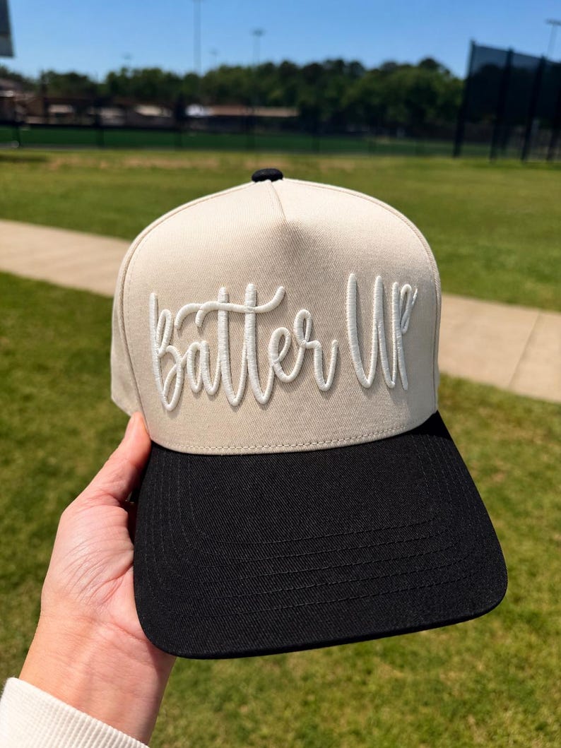 May include: A beige and black baseball cap with the text "Batter Up" embroidered in white script. The cap has a structured front panel and a curved brim. The cap is being held up in front of a green field.