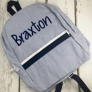 May include: A blue and white striped backpack with the name "Braxton" embroidered in navy blue. The backpack has a navy blue zipper and a white zipper pull. The straps and handle are navy blue.