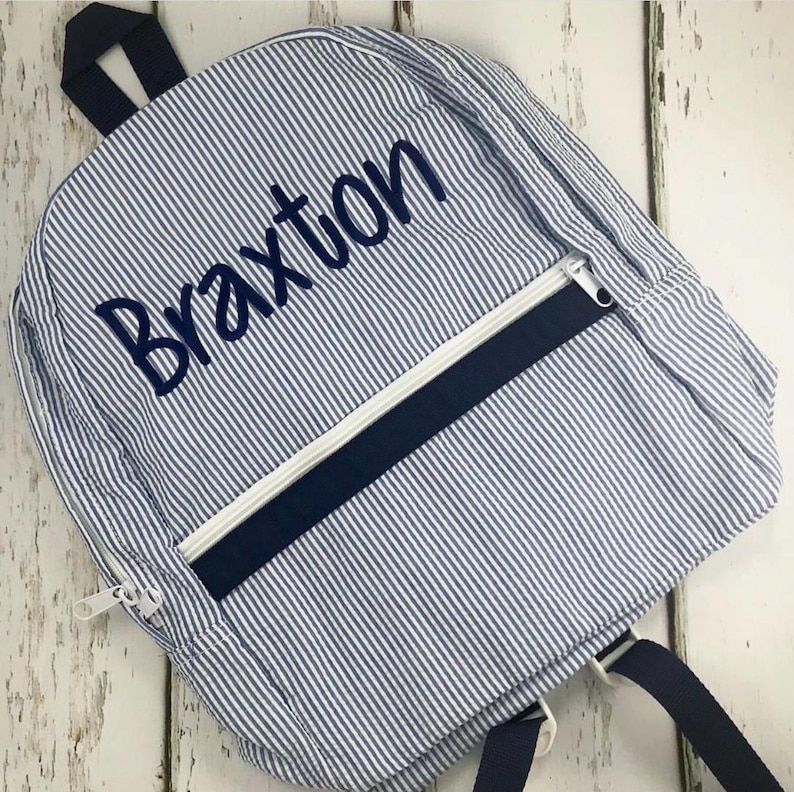 May include: A blue and white striped backpack with the name "Braxton" embroidered in navy blue. The backpack has a navy blue zipper and a white zipper pull. The straps and handle are navy blue.