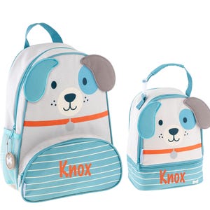 May include: A blue and gray dog-themed backpack and lunch bag set with the name "Knox" printed on both. The backpack has a mesh side pocket and a zippered front pocket. The lunch bag has a top handle and a zippered closure.