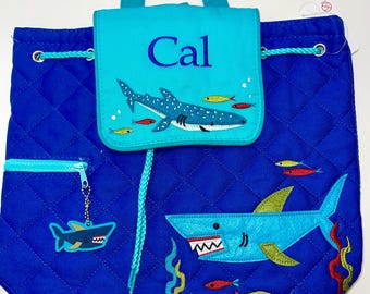 Puede incluir: Una mochila acolchada azul con cierre de cordón. La mochila presenta un diseño de tiburón azul y verde con un pequeño amuleto de tiburón. La solapa delantera tiene un bolsillo turquesa con el nombre "Cal" bordado.