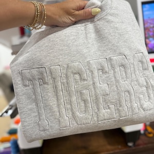 May include: A light gray sweatshirt with the word "TIGERS" embroidered in a lighter gray thread. The sweatshirt is held up by a hand, and the background includes a sewing machine and a monitor.