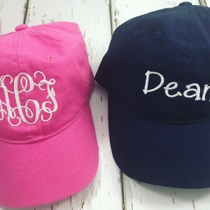 May include: A bright pink baseball cap with white embroidered monogram lettering and a navy blue baseball cap with white embroidered text that reads Dean. Both caps feature a structured crown, a curved brim, and a button on the top center.