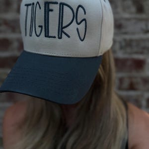 Op de afbeelding: Een beige en marineblauwe baseballcap met het woord "TIGERS" in marineblauw op de voorkant geborduurd. De cap heeft een gebogen klep en een gestructureerde kroon, geschikt voor sportfans.