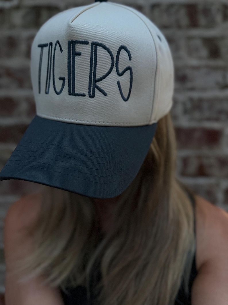 Op de afbeelding: Een beige en marineblauwe baseballcap met het woord "TIGERS" in marineblauw op de voorkant geborduurd. De cap heeft een gebogen klep en een gestructureerde kroon, geschikt voor sportfans.