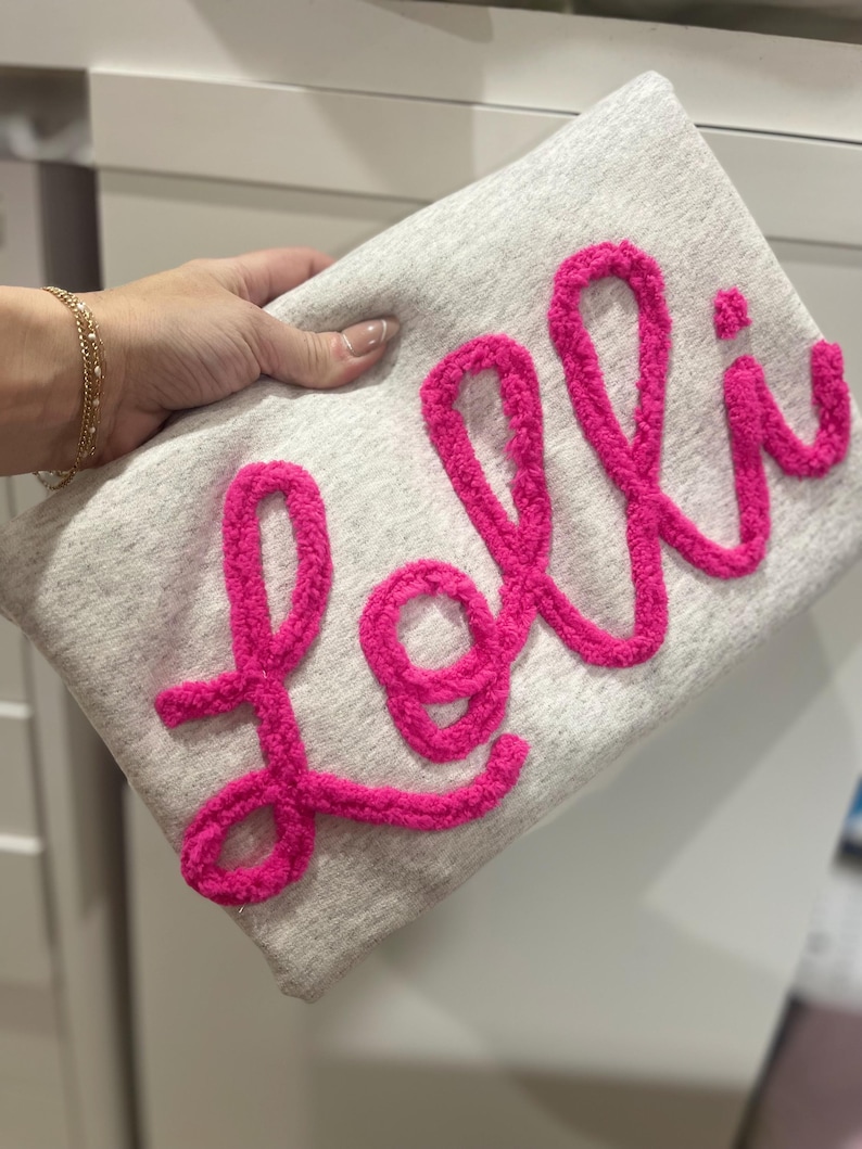 May include: A grey fabric clutch with the name "Lolli" embroidered in pink fluffy yarn.