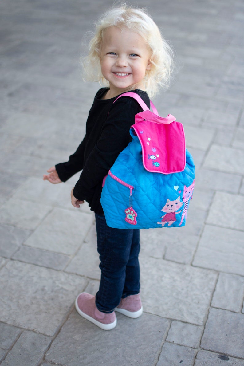 Peut inclure: Un sac &agrave; dos matelass&eacute; bleu avec un rabat et des bretelles roses. Le sac &agrave; dos pr&eacute;sente un motif de chat et une tirette en forme de patte. L'enfant porte un haut noir &agrave; manches longues, un jean bleu et des chaussures roses.