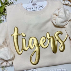May include: A cream-colored sweatshirt with the word "tigers" in gold foil lettering. The sweatshirt is on a white surface with white beads.
