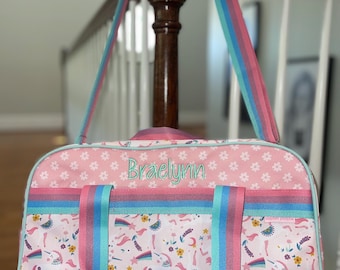 Può includere: Borsa da viaggio rosa con un motivo di unicorni e fiori. La borsa ha una tracolla a righe arcobaleno e il nome "Braelynn" ricamato in verde. La borsa è decorata con un bordo azzurro.