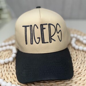 Op de afbeelding: Beige en zwarte baseballcap met het woord "TIGERS" in zwarte letters geborduurd. De cap heeft een gebogen klep en een gestructureerde kroon, geschikt voor vrijetijdskleding. Een modieus accessoire.