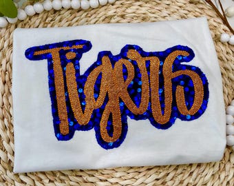 May include: White t-shirt with the word "Tigers" in a gold glitter script font. The lettering is outlined in blue sequins. The shirt is folded and resting on a woven surface.