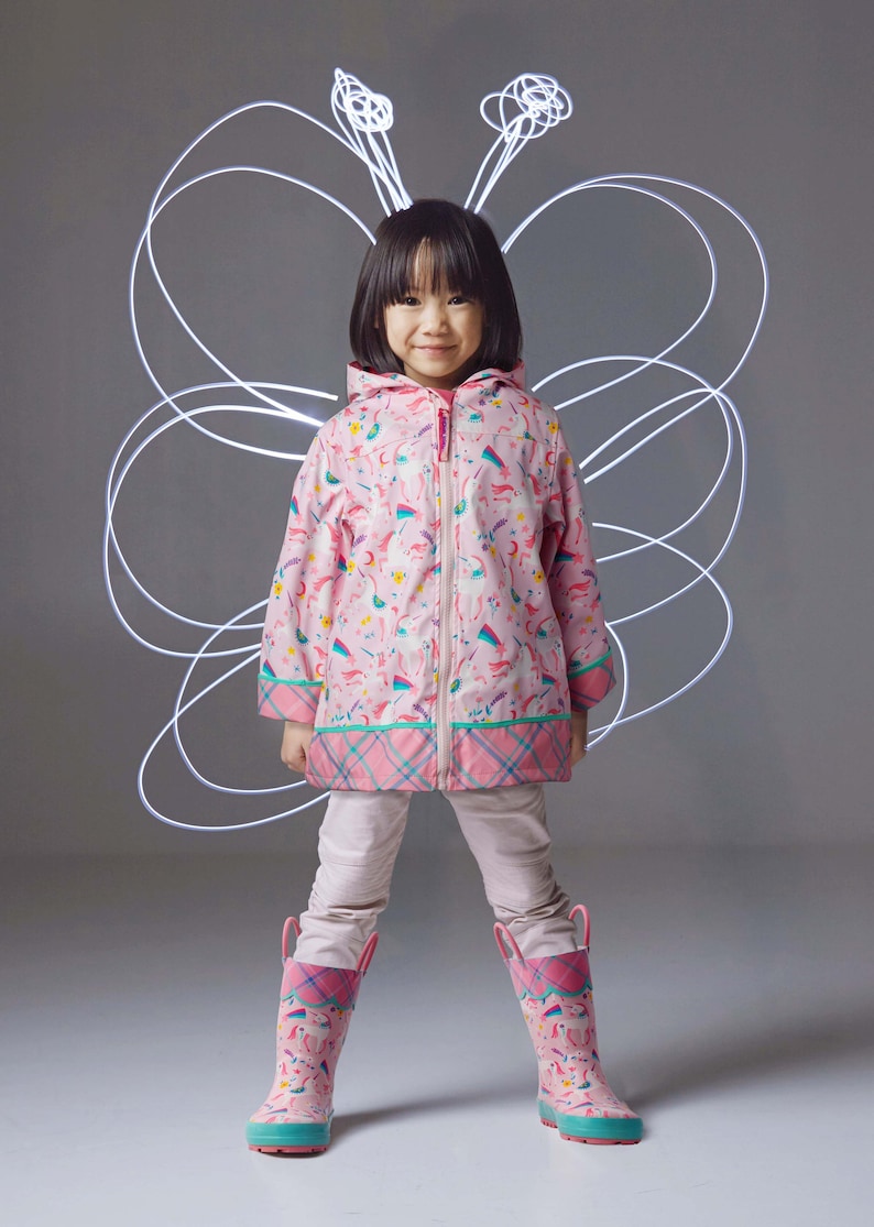 May include: A pink hooded raincoat featuring a repeating pattern of unicorns, rainbows, and stars, paired with matching pink rubber boots with teal soles. The jacket has a front zipper and a plaid trim along the hem and cuffs of the sleeves.