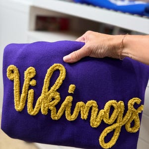 May include: A purple blanket with the word "Vikings" in gold, textured lettering. The blanket is held by a person. The background is blurred, showing a white surface and a blue object.