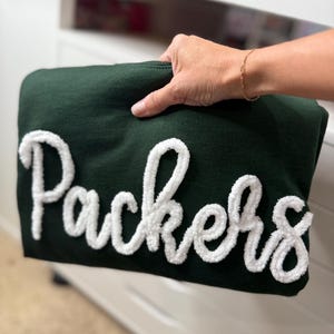 May include: A dark green sweatshirt with the word "Packers" in large, white, fluffy lettering. The sweatshirt is held up by a hand, showcasing the design. The background is a white dresser.