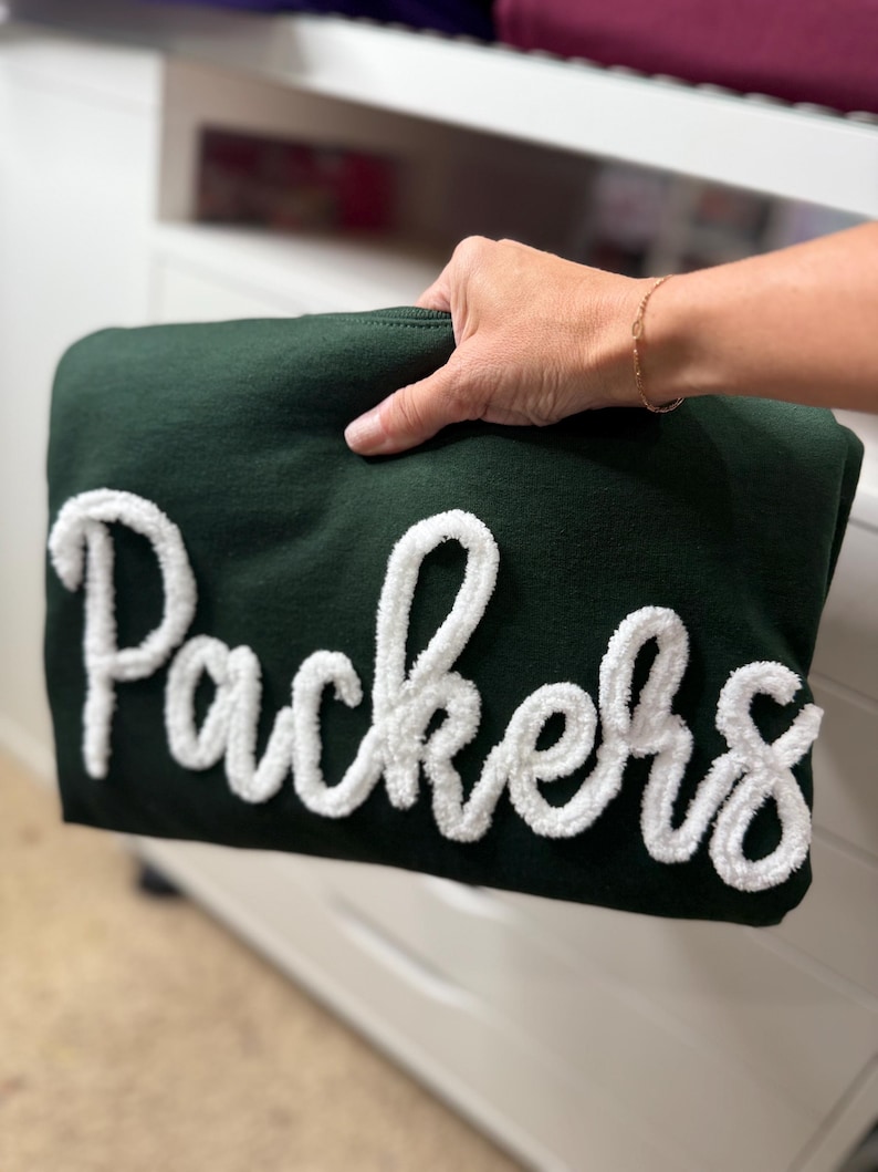 May include: A dark green sweatshirt with the word "Packers" in large, white, fluffy lettering. The sweatshirt is held up by a hand, showcasing the design. The background is a white dresser.