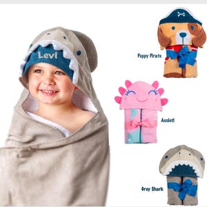 May include: Four hooded towels for babies, featuring a grey shark, a pink axolotl, a grey shark with a blue mouth, and a brown puppy pirate. The towels are rolled up and tied with a ribbon. The towels are personalised with the name "Levi" on the grey shark towel.