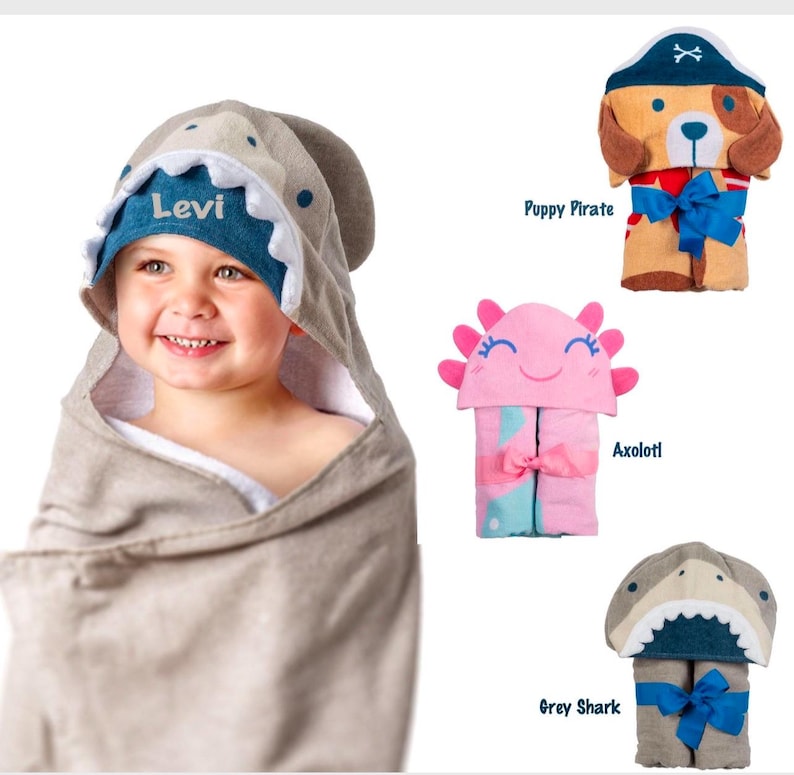 May include: Four hooded towels for babies, featuring a grey shark, a pink axolotl, a grey shark with a blue mouth, and a brown puppy pirate. The towels are rolled up and tied with a ribbon. The towels are personalised with the name "Levi" on the grey shark towel.