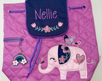 Puede incluir: Una mochila acolchada rosa con cierre de cordón morado y correas moradas. La mochila presenta un elefante bordado con detalles florales y un corazón. La solapa delantera tiene un bolsillo azul marino con el nombre "Nellie" bordado en morado.