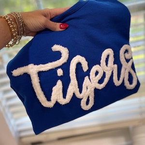 May include: A royal blue sweatshirt with the word "Tigers" in white, textured lettering. The sweatshirt is held up, showcasing the bold text and color contrast. The background is blurred.