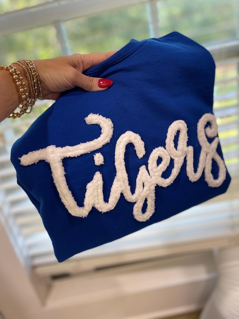May include: A royal blue sweatshirt with the word "Tigers" in white, textured lettering. The sweatshirt is held up, showcasing the bold text and color contrast. The background is blurred.