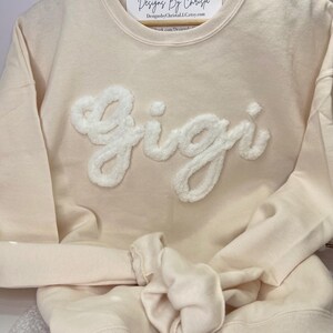 May include: A cream colored sweatshirt with a white fluffy applique of the name "Gigi" on the front. The sweatshirt is folded and hanging on a wooden hanger.