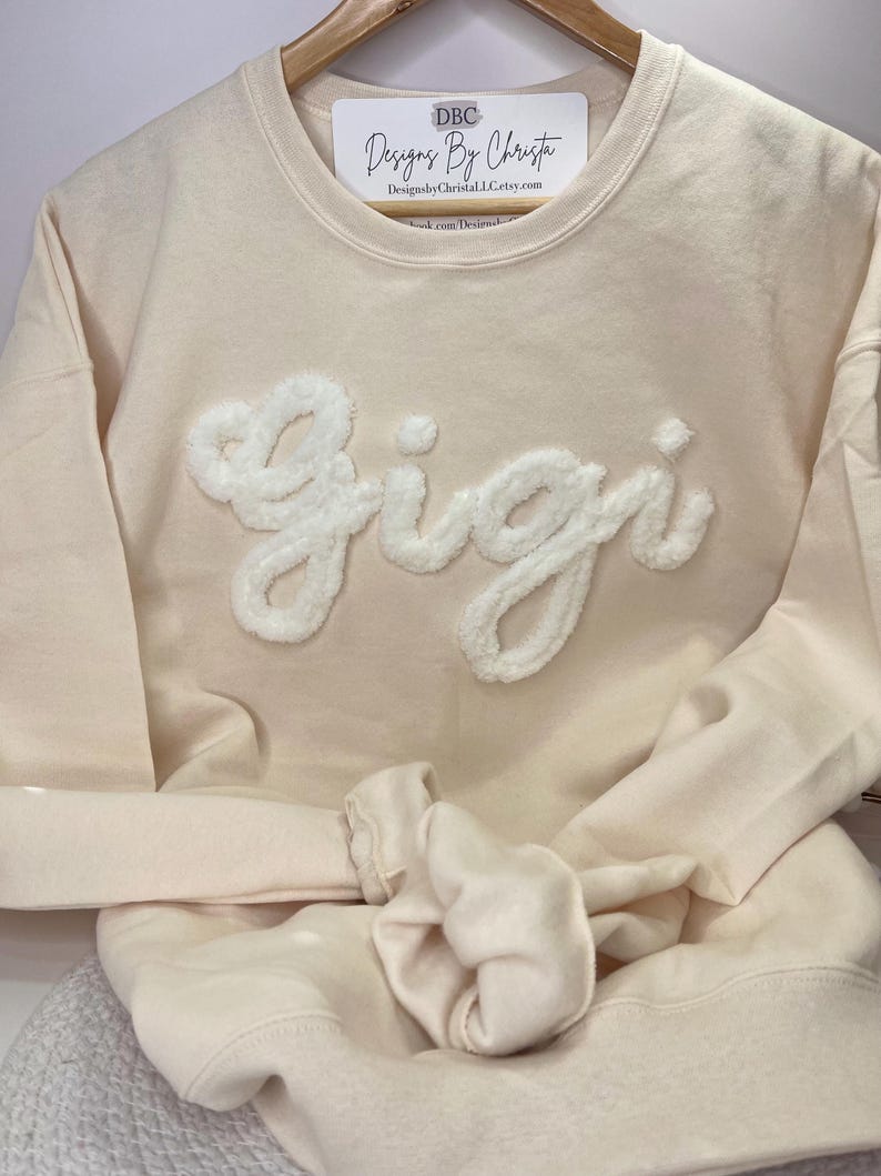May include: A cream colored sweatshirt with a white fluffy applique of the name "Gigi" on the front. The sweatshirt is folded and hanging on a wooden hanger.