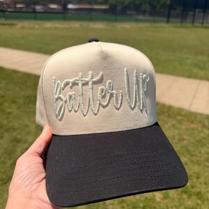 May include: A beige and black baseball cap with the text "Batter Up" embroidered in a cursive font. The cap has a structured front panel and a curved brim. The background shows a green field and a blue sky.