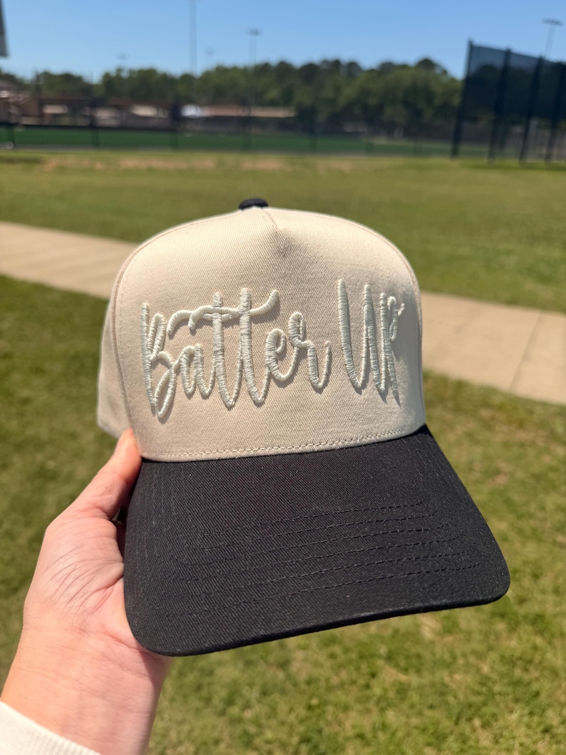 May include: A beige and black baseball cap with the text "Batter Up" embroidered in a cursive font. The cap has a structured front panel and a curved brim. The background shows a green field and a blue sky.