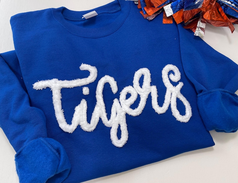 May include: Royal blue sweatshirt with the word "Tigers" in white, textured lettering across the front. The sweatshirt has a crew neck and ribbed cuffs. A red, white, and blue pom-pom is visible in the upper right corner.