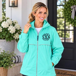 May include: A bright turquoise hooded windbreaker jacket featuring a full-length silver zipper, long sleeves with adjustable cuffs, and a black embroidered monogram on the left chest. The jacket includes a horizontal reflective silver stripe.