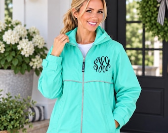 May include: A bright turquoise hooded windbreaker jacket featuring a full-length silver zipper, long sleeves with adjustable cuffs, and a black embroidered monogram on the left chest. The jacket includes a horizontal reflective silver stripe.
