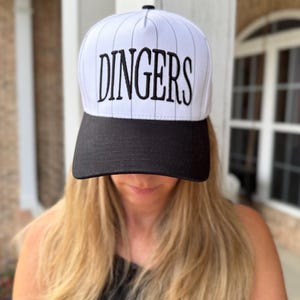 May include: A white and black baseball cap with the word "DINGERS" embroidered in black. The cap has a black brim and a white crown with vertical pinstripes. The cap is worn by a person with blonde hair.
