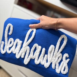 May include: A folded blue blanket with the word "Seahawks" in white, fluffy lettering. The blanket is held by a person's hand, and the background is a white dresser.
