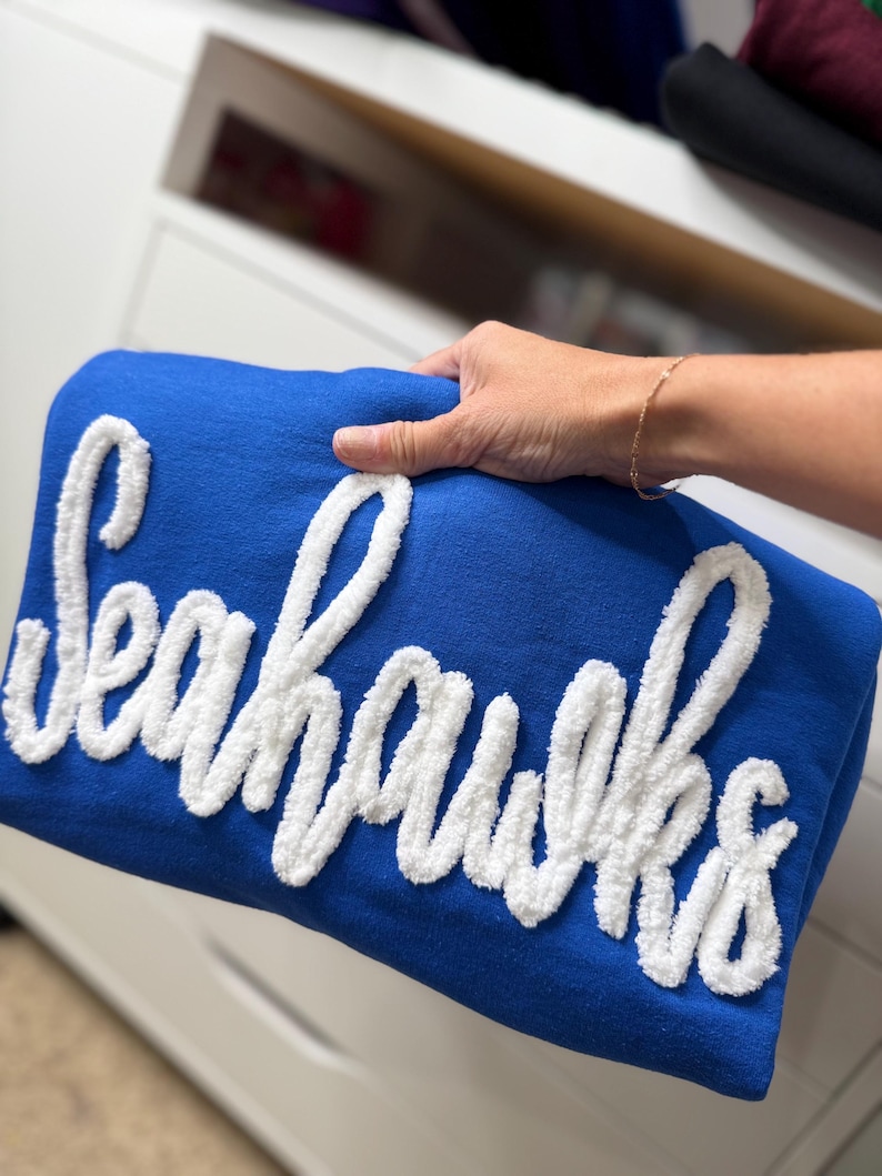 May include: A folded blue blanket with the word "Seahawks" in white, fluffy lettering. The blanket is held by a person's hand, and the background is a white dresser.