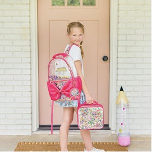 May include: A clear backpack with a large pink bow and colorful confetti-filled pockets, paired with a matching rectangular lunch bag. The backpack features a small attached coin purse and pink trim, while the lunch bag has a matching confetti design.