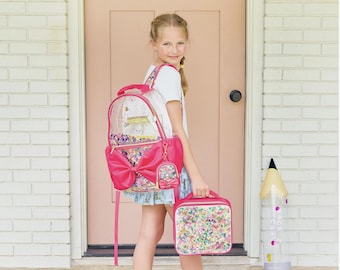 May include: A clear backpack with a large pink bow and colorful confetti-filled pockets, paired with a matching rectangular lunch bag. The backpack features a small attached coin purse and pink trim, while the lunch bag has a matching confetti design.