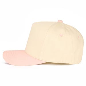 Op de afbeelding: Een baseballcap in cr&egrave;me en roze. De kroon is cr&egrave;me en de klep is roze. De cap heeft een gestructureerd ontwerp met een platte klep en een roze knoop. Een modieus accessoire voor dagelijks gebruik.