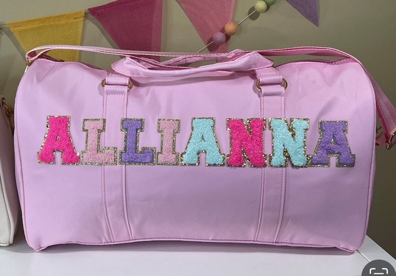 May include: A light pink duffel bag with the name "ALLIANNA" spelled out in colorful, sparkly letters.
