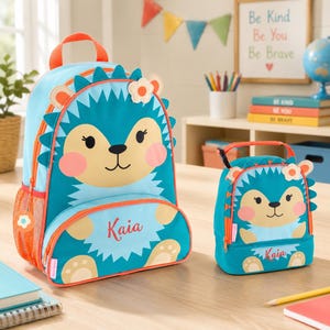 May include: A teal and orange hedgehog-themed backpack and matching lunch bag. The backpack features a large hedgehog face with the name "Kaia" in red script. The lunch bag has a similar design. Both items have orange accents and are designed for children.