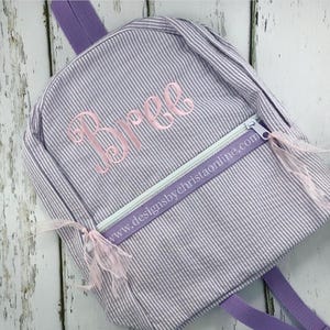 May include: A small, seersucker-striped backpack in shades of lavender and white. The backpack features a front zippered pocket with a light green zipper and a pink embroidered name, "Bree". The website address "www.designsbychristaonline.com" is printed on the zipper pull.