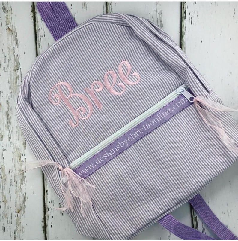 May include: A small, seersucker-striped backpack in shades of lavender and white. The backpack features a front zippered pocket with a light green zipper and a pink embroidered name, "Bree". The website address "www.designsbychristaonline.com" is printed on the zipper pull.
