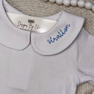 May include: White short-sleeved onesie with a Peter Pan collar. The collar is embroidered with the name "stratton" in blue thread. A tag with the text "DBC Designs By Christi" is attached to the collar.