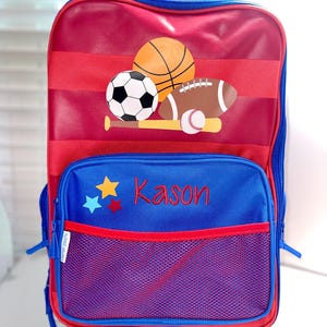 May include: Red and blue rolling backpack with a sports theme. The front features a basketball, football, baseball, and a bat. The backpack has a mesh pocket and the name "Kason" is embroidered on the front.