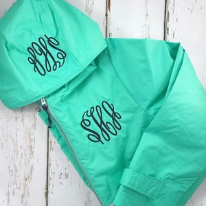 May include: A turquoise hooded jacket with a black monogram on the front and hood. The jacket has a zipper closure and a drawstring hood.