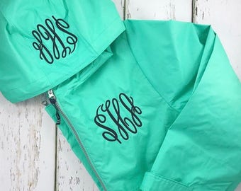 May include: A turquoise hooded jacket with a black monogram on the front and hood. The jacket has a zipper closure and a drawstring hood.