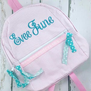 May include: Pink and white striped backpack with the name "Evee June" embroidered in teal. The backpack has a front zipper pocket with a pink ribbon and teal polka dot ribbons attached. The straps and handle are pink.