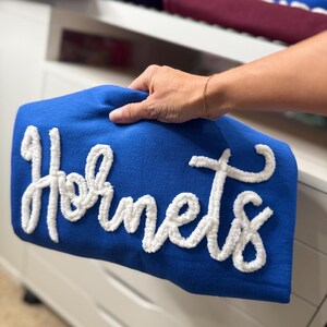 May include: A royal blue pillow with the word "Hornets" in white, textured, cursive lettering. The pillow is held by a person's hand. Other folded items are visible in the background.