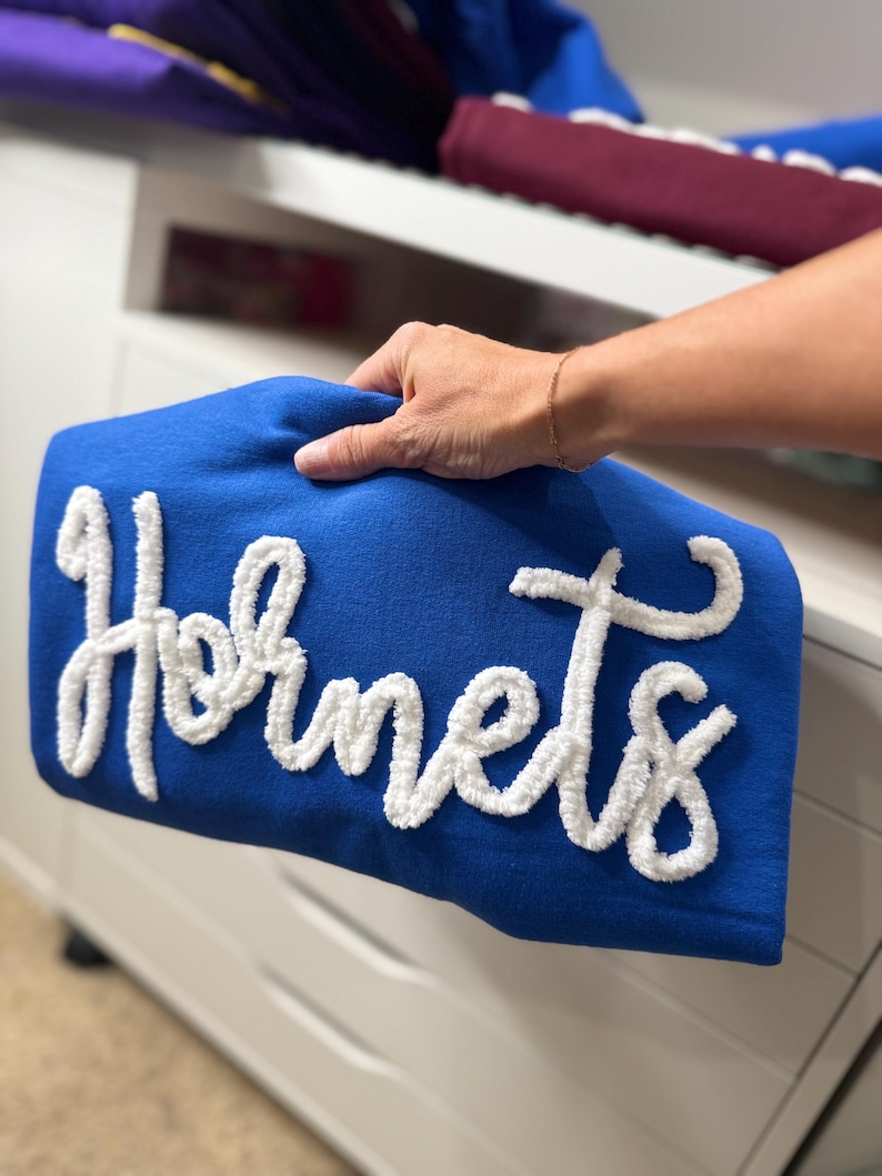 May include: A royal blue pillow with the word "Hornets" in white, textured, cursive lettering. The pillow is held by a person's hand. Other folded items are visible in the background.
