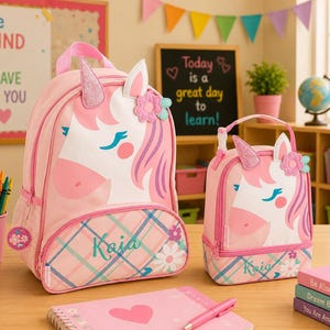 Puede incluir: Mochila rosa de unicornio y bolsa de almuerzo a juego. La mochila y la bolsa de almuerzo presentan un diseño de cara de unicornio con cuerno, orejas y detalles florales. La mochila tiene un estampado de cuadros en la parte inferior con el nombre "Kaia".