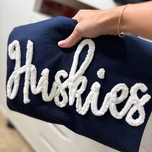 May include: A navy blue sweatshirt with the word "Huskies" in large, white, textured lettering. The sweatshirt is held up, showcasing the design. The background is blurred, with other folded garments visible.